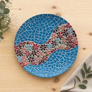Peut inclure: Une assiette décorative ronde avec un motif de mosaïque. L'assiette présente un fond bleu avec un motif de mosaïque et un motif floral au centre. Les fleurs sont dans les tons de rouge, rose, vert et marron. L'assiette est posée sur une surface en bois.