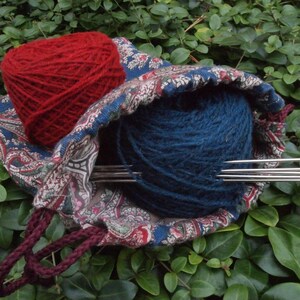Pu&ograve; includere: Una borsa in tessuto con motivo paisley contenente un gomitolo di lana rosso e uno blu. La borsa ha un cordoncino di chiusura color marrone e ci sono ferri da maglia che sporgono dalla borsa.