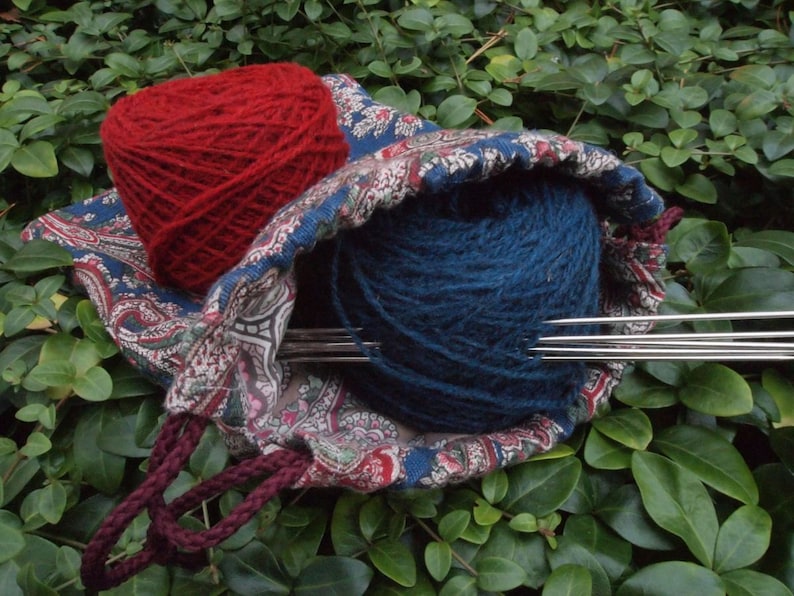 Pu&ograve; includere: Una borsa in tessuto con motivo paisley contenente un gomitolo di lana rosso e uno blu. La borsa ha un cordoncino di chiusura color marrone e ci sono ferri da maglia che sporgono dalla borsa.