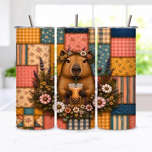 Könnte beinhalten: Ein zylindrischer Becher mit einem Patchwork-Muster in Blau-, Orange- und Rosatönen. Das Design zeigt ein zentrales Wasserschwein mit Blumenkranz, das Bubble Tea trinkt, umgeben von Lavendel- und Gänseblümchen-Blumenarrangements.