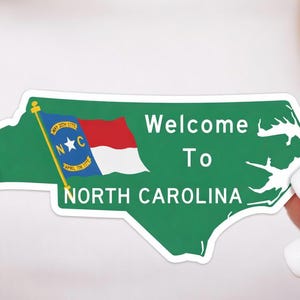 K&ouml;nnte beinhalten: Ein gr&uuml;ner Aufkleber in Form von North Carolina mit den Worten "Welcome To North Carolina". Die Staatsflagge ist links abgebildet. Der Aufkleber wird von einer Person mit manik&uuml;rten N&auml;geln gehalten.