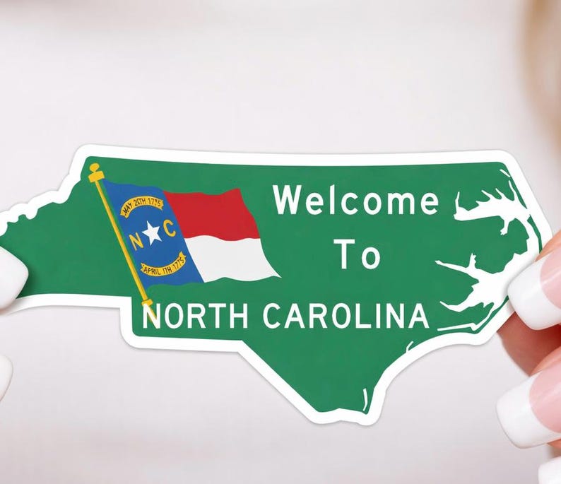K&ouml;nnte beinhalten: Ein gr&uuml;ner Aufkleber in Form von North Carolina mit den Worten "Welcome To North Carolina". Die Staatsflagge ist links abgebildet. Der Aufkleber wird von einer Person mit manik&uuml;rten N&auml;geln gehalten.