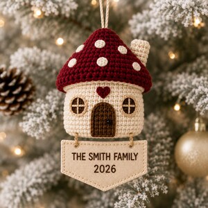 Puede incluir: Un adorno de ganchillo con forma de casa seta, con sombrero rojo oscuro y puntos blancos, base color crema, puerta marr&oacute;n y un peque&ntilde;o coraz&oacute;n rojo. Debajo cuelga una etiqueta de tela beige con el texto THE SMITH FAMILY 2026 en negro.