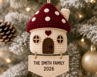 May include: A crochet mushroom house ornament with a dark red cap featuring white spots, a cream-colored base, a brown door, and a small red heart. A beige fabric sign hangs below with the text THE SMITH FAMILY 2026 printed in black ink.