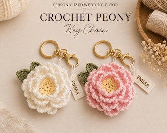 May include: Two crochet peony key chains, one white and one pink, each with a yellow center, green leaves, a gold-tone metal ring, and a rectangular tag printed with the name EMMA. Text above reads PERSONALIZED WEDDING FAVOR CROCHET PEONY Key Chain.