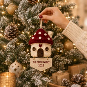 Puede incluir: Un adorno de casa de hongo tejido a ganchillo con techo rojo oscuro y puntos blancos, base color crema, puerta marr&oacute;n oscuro y un peque&ntilde;o coraz&oacute;n sobre la entrada. Una etiqueta de tela beige cuelga debajo con el texto THE SMITH FAMILY 2026.