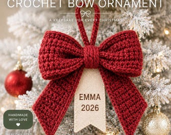 May include: A deep red crochet bow ornament featuring a central loop for hanging and a beige fabric tag hanging from the center. The tag is printed with the name EMMA and the year 2026. The bow has a textured, hand-stitched appearance.
