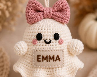 May include: A cream-colored crocheted ghost ornament featuring a pink crocheted bow on top, black embroidered eyes and a smile, pink cheeks, and a rectangular fabric patch on the front with the name EMMA stitched in dark brown thread.