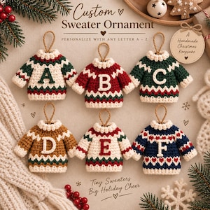 May include: Six crocheted sweater ornaments with hanging loops, each featuring a unique letter from A to F. The sweaters display various color combinations including cream, red, green, brown, and navy blue with geometric patterns and text.