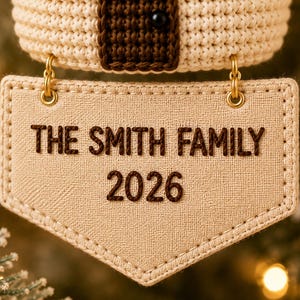 Puede incluir: Un adorno de tela beige con forma de escudo, que presenta el texto bordado THE SMITH FAMILY 2026 en marr&oacute;n oscuro. El adorno est&aacute; sujeto por dos anillos de metal dorado a una secci&oacute;n superior de ganchillo en color crema y marr&oacute;n con una peque&ntilde;a cuenta negra.