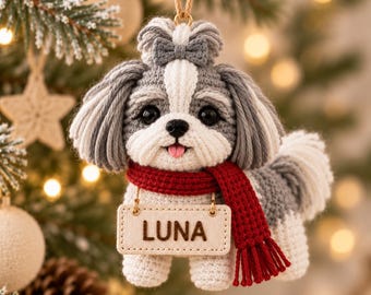 May include: A crocheted Shih Tzu dog ornament featuring gray and white yarn, a small gray bow on its head, and a red scarf. A rectangular beige tag hangs from the scarf with the name LUNA printed in dark brown letters. The ornament hangs by a string.