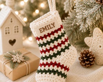 May include: A crocheted Christmas stocking featuring a cream, dark green, and red striped pattern. The stocking has a cream-colored cuff with a rectangular fabric tag sewn onto the front, displaying the name EMILY in brown capital letters.