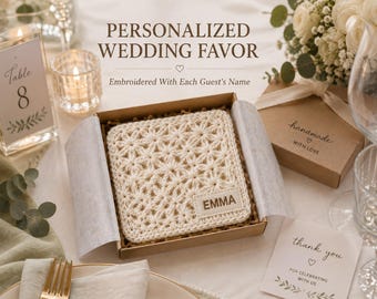 May include: A square, off-white crocheted coaster with a star-like pattern, featuring a rectangular fabric tag embroidered with the name EMMA. The coaster is presented inside an open brown cardboard gift box with light-colored tissue paper.