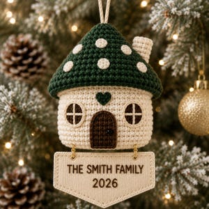 Puede incluir: Un adorno de casa de ganchillo con techo verde oscuro y lunares blancos, cuerpo color crema, puerta marr&oacute;n oscuro, dos ventanas y un peque&ntilde;o coraz&oacute;n. Debajo cuelga un cartel rectangular beige con el texto THE SMITH FAMILY 2026.