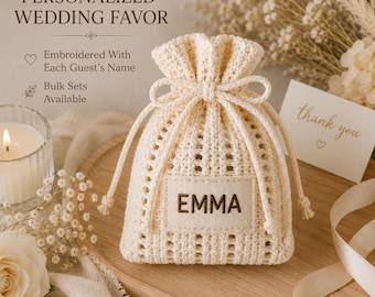 May include: A cream-colored crocheted drawstring pouch featuring a rectangular fabric label with the name EMMA embroidered in brown. The pouch includes a matching drawstring closure and is displayed with text reading PERSONALIZED WEDDING FAVOR.
