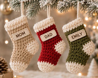 May include: Three crocheted Christmas stockings hanging from a pine branch. The left stocking is beige, the center is red, and the right is olive green. Each features a white cuff, heel, and toe, with a rectangular wooden tag labeled MOM, DAD, or EMILY.