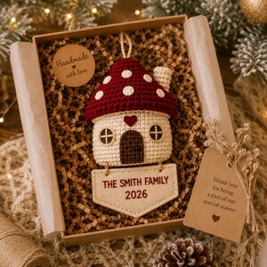 Puede incluir: Un adorno de casa de hongo tejido a ganchillo con techo rojo de lunares blancos, base color crema y puerta marr&oacute;n oscuro. Una etiqueta de tela beige unida en la parte inferior muestra el texto bordado THE SMITH FAMILY 2026 en rojo oscuro.
