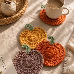 May include: A set of three round, crocheted pumpkin-shaped coasters in mustard yellow, burnt orange, and muted purple, each featuring a small, textured green stem at the top. The coasters are arranged on a flat surface, with one supporting a ceramic mug.