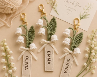May include: Three crochet lily of the valley keychains with gold-tone lobster clasps. Each features a green stem, white bell-shaped flowers, and a beige fabric tag embroidered with the names Olivia, Emma, or Sophia, tied with a small ribbon.