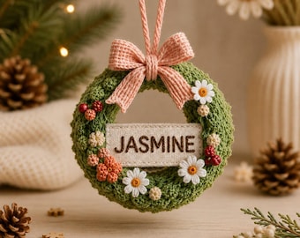 May include: A circular green crocheted wreath ornament featuring a light pink fabric bow at the top and a rectangular center tag embroidered with the name JASMINE. The wreath is decorated with small white daisy flowers and clusters of red berries.