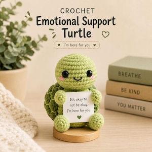 Op de afbeelding: Een lichtgroen gehaakt schildpadfiguurtje met zwarte ogen en roze wangen, dat een wit kaartje vasthoudt met de tekst: It's okay to not be okay. I'm here for you. De schildpad staat op een ronde houten voet, met daarboven de tekst: CROCHET Emotional Support Turtle.