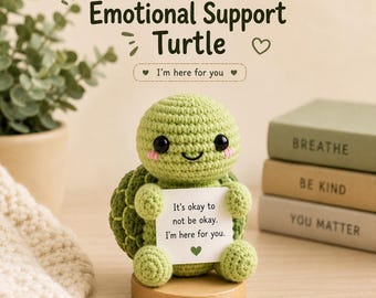 May include: A light green crocheted turtle figurine with black eyes and pink cheeks, holding a white card that reads: It's okay to not be okay. I'm here for you. The turtle sits on a round wooden base, with text above reading: CROCHET Emotional Support Turtle.