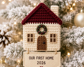 May include: A crocheted house ornament with a dark red roof, cream walls, a brown door, and a green wreath. A beige fabric banner hangs below with the text "OUR FIRST HOME 2026" and a small red heart. The ornament hangs from a white string.