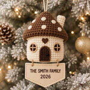 Puede incluir: Un adorno de ganchillo con forma de casa seta, con techo marr&oacute;n de lunares blancos, cuerpo color crema, puerta marr&oacute;n oscuro y un peque&ntilde;o coraz&oacute;n. Debajo cuelga una placa de tela beige con el texto THE SMITH FAMILY 2026.