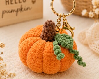 May include: A crocheted orange pumpkin keychain featuring a brown textured stem and a green curly vine. The pumpkin is attached to a gold-toned metal split ring and a lobster clasp. The design is soft and rounded with visible yarn stitches.
