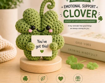 May include: A green crocheted four-leaf clover figure with a curled stem, black bead eyes, and pink stitched cheeks. It holds a white square sign with the text 'You've got this!' and a small green heart. The figure sits on a round wooden base.
