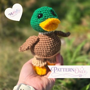 May include: A crocheted mallard duck toy featuring a green head, a bright yellow beak, black bead eyes, and a brown body with light tan feet. The image includes a white heart-shaped logo with the text MohyKi and a banner stating Pattern Only.