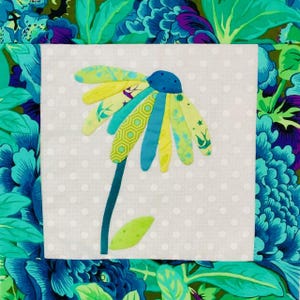 Könnte beinhalten: Ein Stoffquadrat mit einer applizierten Blume mit gelben, türkisfarbenen und blauen Blütenblättern. Die Blume hat eine blaue Mitte und einen türkisfarbenen Stiel mit einem grünen Blatt. Der Hintergrund ist weiß mit kleinen grauen Punkten, umgeben von einem floralen Stoffrand.