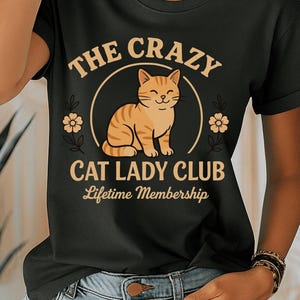 Peut inclure: T-shirt noir avec l'inscription "THE CRAZY CAT LADY CLUB Lifetime Membership". Le motif représente un chat tigré orange souriant à l'intérieur d'un cercle, avec des motifs floraux. Le t-shirt est de style col rond.