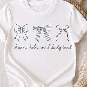 Peut inclure: T-shirt blanc avec trois illustrations de nœuds noirs et le texte "chosen. holy. and dearly loved." Les nœuds sont de styles différents. Le t-shirt est fait d'une matière douce.