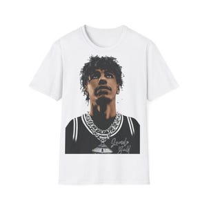 以下が含まれることがあります： 白いTシャツ。カールした髪の人物の肖像画が描かれており、白黒のジャージとシルバーのチェーンを着用しています。「Jarvis Golf」の文字が下にイタリック体で印刷されています。