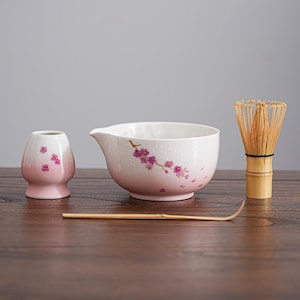 Puede incluir: Un juego de té matcha de cerámica blanca con detalles florales rosas. El juego incluye un cuenco, un batidor, una cuchara y un recipiente pequeño. El batidor es de bambú. El juego está dispuesto sobre una superficie de madera.