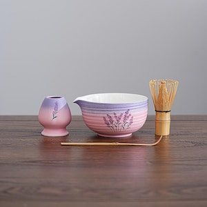 May include: A matcha tea set with a lavender theme. The set includes a pink and purple bowl, a small vase, a bamboo whisk, and a bamboo scoop. The bowl and vase have a lavender floral design.