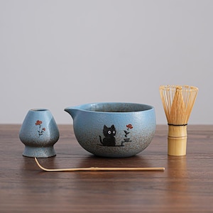 May include: A blue ceramic matcha tea set featuring a black cat design. The set includes a bowl, whisk, small vase, and bamboo scoop. The bowl and vase have a floral design. The whisk is made of bamboo.