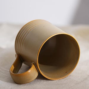 Puede incluir: Una taza de cer&aacute;mica con exterior beige y un interior marr&oacute;n. La taza tiene un asa redondeada y estr&iacute;as horizontales alrededor del cuerpo. La taza est&aacute; inclinada, revelando el interior y el asa.