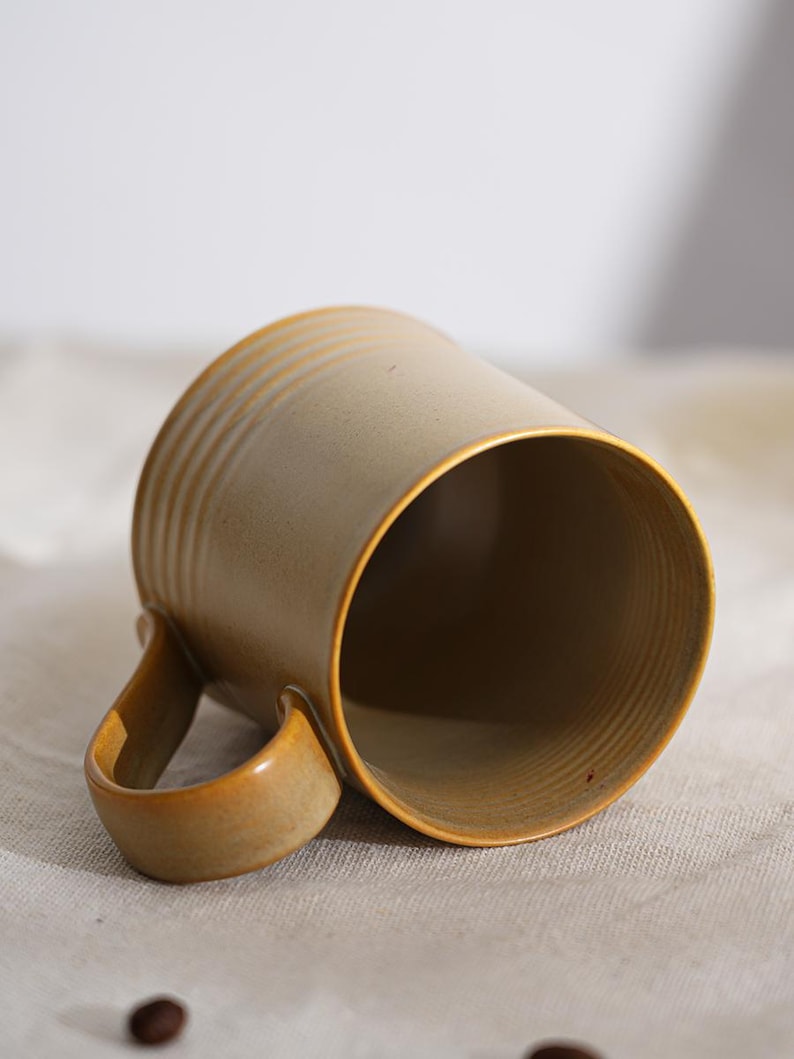 Puede incluir: Una taza de cer&aacute;mica con exterior beige y un interior marr&oacute;n. La taza tiene un asa redondeada y estr&iacute;as horizontales alrededor del cuerpo. La taza est&aacute; inclinada, revelando el interior y el asa.