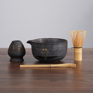 Puede incluir: Un juego de té matcha de cerámica negra con ilustraciones de gatos dorados. El juego incluye un cuenco, un batidor, una cuchara y un pequeño jarrón con diseños de corazón. El batidor es de bambú. Los artículos están dispuestos sobre una superficie de madera.