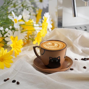May include: A brown ceramic coffee cup and saucer set with a black cat design. The cup contains a latte with a heart-shaped foam design. The set sits on a white cloth, with coffee beans scattered nearby.