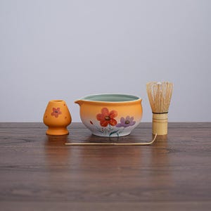 May include: A matcha tea set featuring a ceramic bowl with a floral design, a bamboo whisk, a small ceramic holder, and a bamboo scoop. The bowl is orange with red and purple flowers. The whisk has a bamboo handle. The set is on a wooden surface.