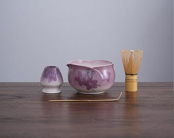 May include: A matcha tea set featuring a lavender-colored ceramic bowl with floral designs, a matching whisk holder, a bamboo whisk, and a bamboo scoop. The set is arranged on a dark wooden surface.
