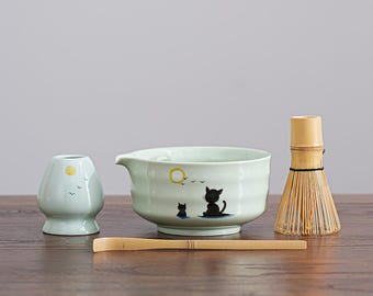 May include: A matcha tea set featuring a light green ceramic bowl with a cat illustration, a whisk, a scoop, and a small ceramic holder. The bowl has a spout for pouring. The set is arranged on a wooden surface.