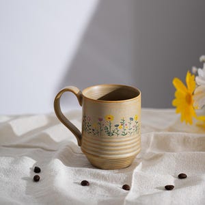 May include: A beige ceramic mug with a floral design featuring yellow, blue, and pink flowers. The mug has a curved handle and horizontal ridges around the base. Coffee beans are scattered around the mug on a white cloth.