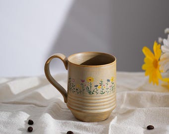 Puede incluir: Taza de cerámica beige con un diseño floral con flores amarillas, azules y rosas. La taza tiene un asa curva y estrías horizontales alrededor de la base. Granos de café esparcidos.