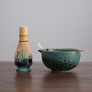 Puede incluir: Un juego de té matcha con un cuenco de cerámica verde con boquilla, un batidor de bambú y una cuchara de bambú. El cuenco tiene pinceladas negras y huellas de patas. El batidor es de bambú.