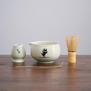 May include: A matcha tea set with a light green ceramic bowl featuring a black cat design, a whisk, a small holder with paw prints, and a bamboo scoop. The set is on a wooden surface.