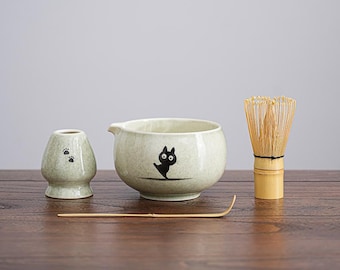 May include: A matcha tea set with a light green ceramic bowl featuring a black cat design, a whisk, a small holder with paw prints, and a bamboo scoop. The set is on a wooden surface.